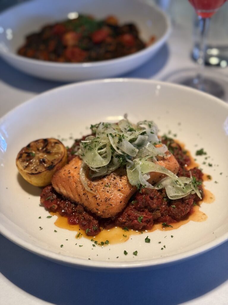 Seared salmon on tomato sauce, topped with shaved fennel and herbs, served with grilled lemon on a white plate.