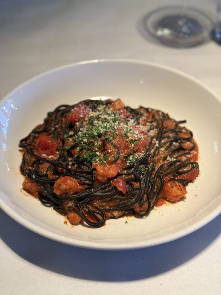 A bowl of black pasta with tomato sauce, shrimp, and grated cheese, served on a white plate.