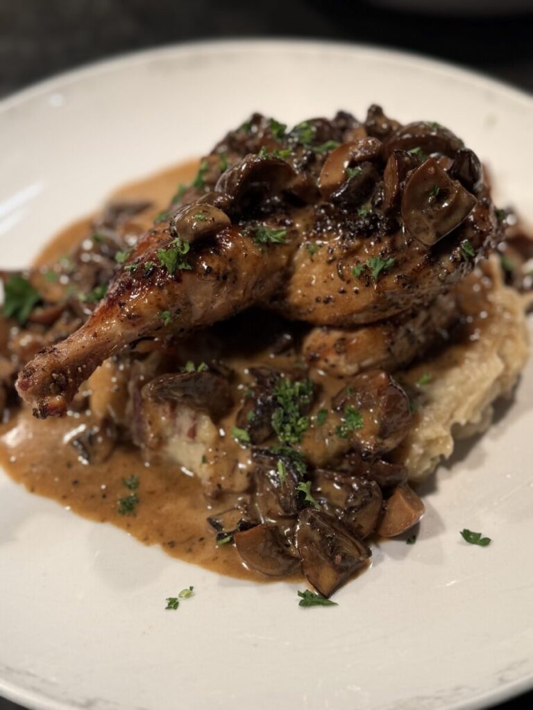 Roast chicken served on mashed potatoes, topped with mushroom gravy and garnished with chopped parsley on a white plate.