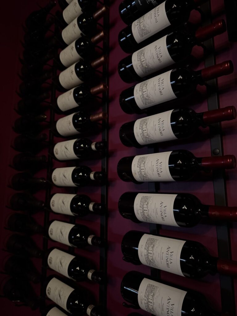 Rows of red wine bottles with white labels stored horizontally on a wall-mounted rack against a dark background.