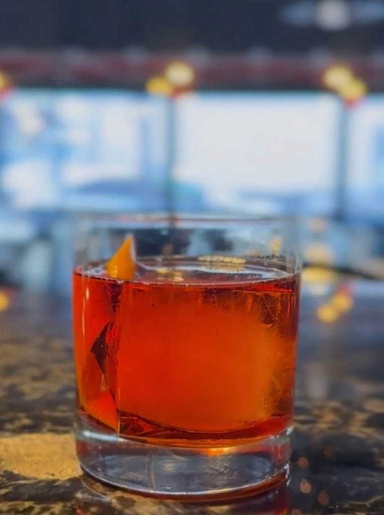 A glass of amber cocktail with ice and an orange peel garnish sits on a reflective surface, blurred background.